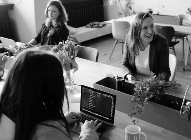 Photo of three people sitting in a room, with one of them using a laptop and another one is smiling